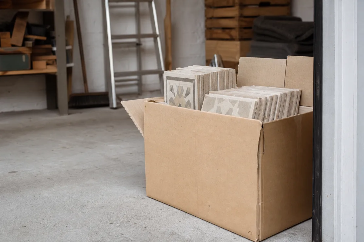 Pile de carreaux de rechange restants soigneusement rangés dans un carton posé au sol d’un garage